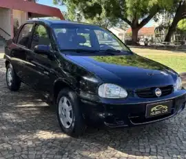 Vista de três quartos dianteira de um sedã Chevrolet preto estacionado sobre uma superfície de pedra com padrão, tendo árvores e um prédio rosa ao fundo.