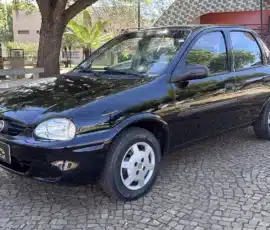 Vista de três quartos dianteira de um sedã Chevrolet preto estacionado sobre uma superfície de pedra com padrão, tendo árvores e um prédio rosa ao fundo.