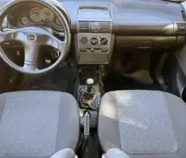 Vista interna da parte dianteira da cabine de um carro: volante, painel, console central e alavanca de câmbio manual entre dois bancos de tecido.