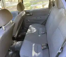 Banco traseiro de um carro compacto com estofamento cinza estampado e cintos de segurança visíveis; vista de um parque pela janela.
