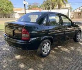 Sedã preto de quatro portas estacionado em uma rua de paralelepípedos com árvores acima, dia ensolarado.