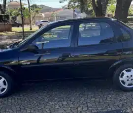 Sedã preto de quatro portas estacionado em uma rua de paralelepípedos em uma área residencial ensolarada, com árvores e casas ao fundo.