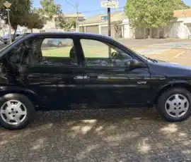 Sedã preto de quatro portas estacionado em uma rua de paralelepípedos em uma área residencial ensolarada, com árvores e casas ao fundo.