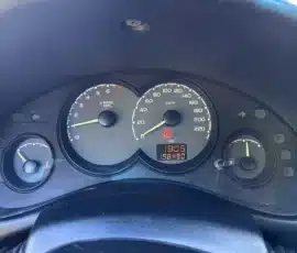 Painel de instrumentos do carro: velocímetro até 220 km/h e conta-giros até 7.000 rpm, com relógio digital marcando 18:05 e hodômetro indicando 198.490 km; luz de advertência do freio de estacionamento (vermelha) acesa; indicadores de nível de combustível e de temperatura visíveis.
