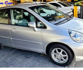 Silver Honda minivan parked at a dealership with other cars in the background, side view visible.