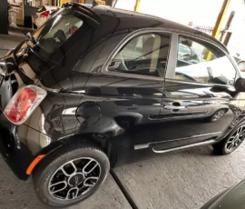 Black compact car (Fiat 500) parked diagonally in a showroom, front to the right, with glossy finish and stylish alloy wheels