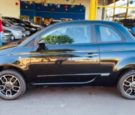 Black Fiat 500 two-door car side profile parked on a street in front of a dealership with yellow-framed windows; motorcycle showroom in background visible in reflection on car surface