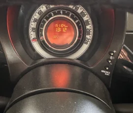 Close-up of a car instrument cluster with a circular speedometer and red digital display showing 15104 km, time 13:12, and 32°C; steering wheel in foreground.