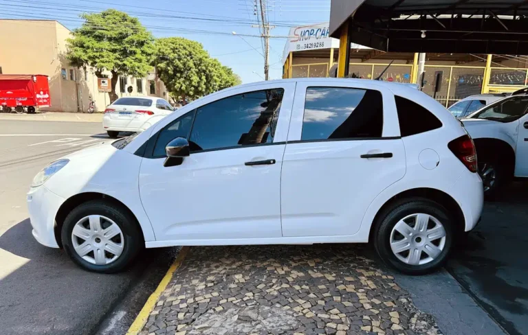 Side view of a white hatchback car parked along a curb on a sunny urban street with buildings in the background.