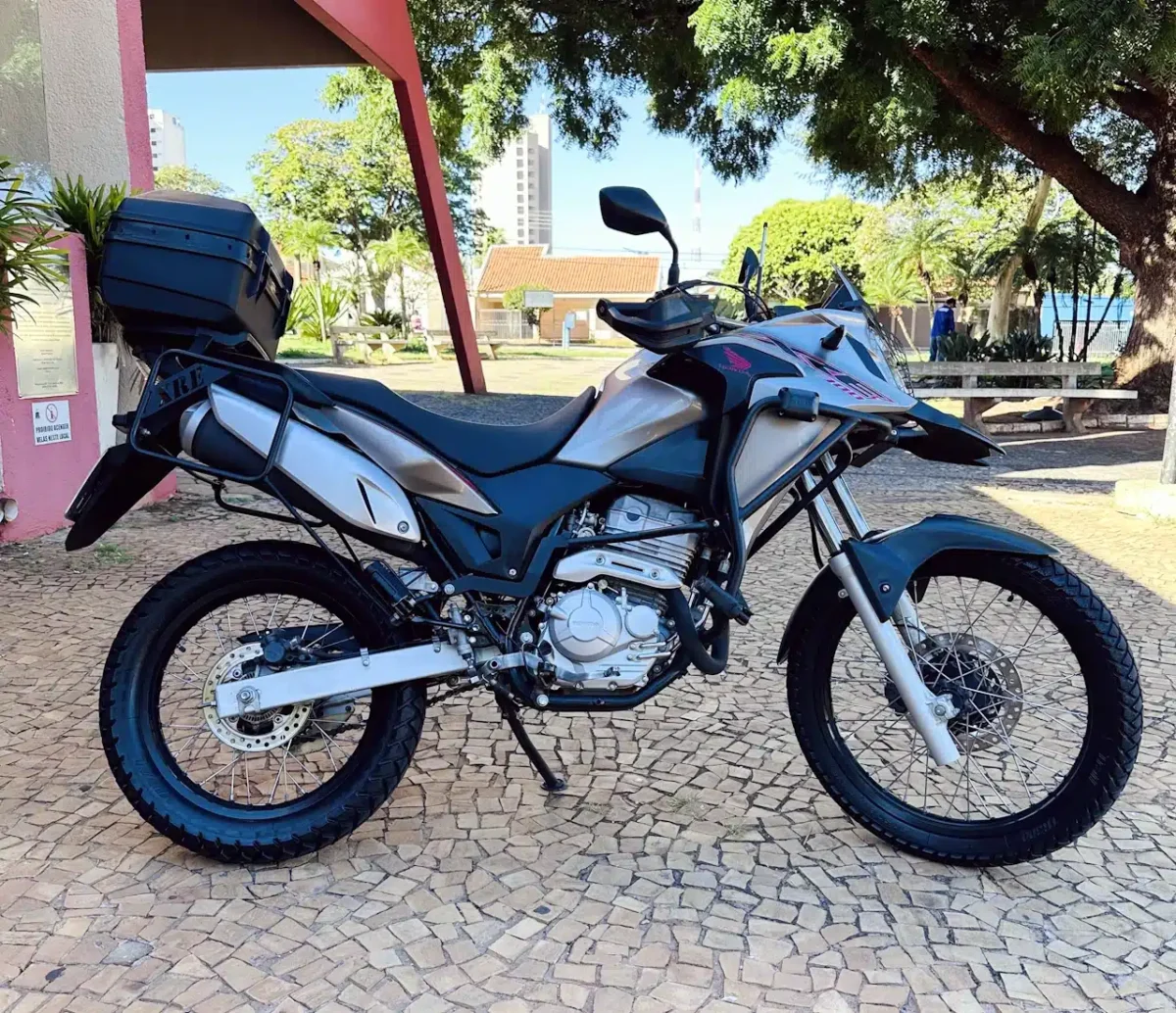 Adventure motorcycle with a top case and side panniers parked on a stone-paved area, trees and benches in the background.