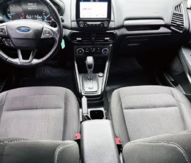 Front interior of a Ford car showing steering wheel, center touchscreen, and gear shifter between two gray seats.