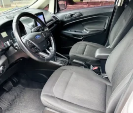 Front interior view of a Ford car showing the steering wheel, dashboard screen, and grey fabric seats with rubber floor mats in the foreground.