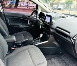 Front interior of a car showing the steering wheel, center touchscreen, gear shifter, and two black fabric seats with a view of the street outside the windows.