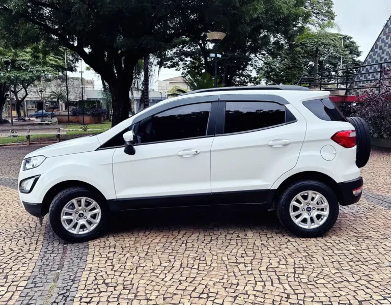 White compact SUV parked on a tan mosaic pavement with trees and buildings in the background (side view).