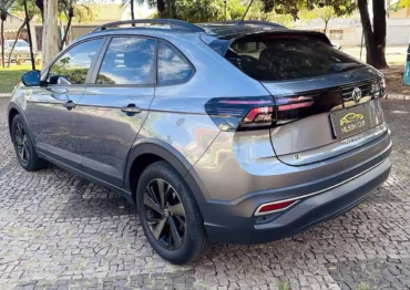 Rear three-quarter view of a metallic gray Volkswagen Nivus SUV parked on a cobblestone lot with roof rails and black alloy wheels on a sunny day, reflecting surroundings.
