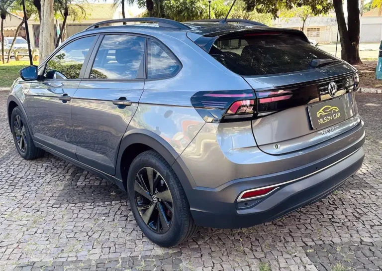 Rear three-quarter view of a metallic gray Volkswagen Nivus SUV parked on a cobblestone lot with roof rails and black alloy wheels on a sunny day, reflecting surroundings.