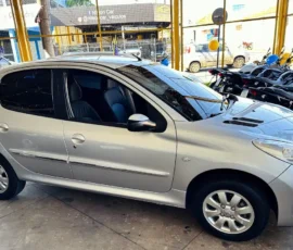 Three-quarter view of a silver compact hatchback parked inside a showroom, with motorcycles behind a yellow fence in the background.