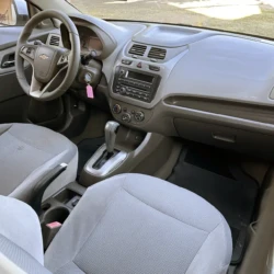 Interior dianteiro de um carro Chevrolet cinza: volante, painel, console central e alavanca de câmbio visíveis, com as chaves na ignição.