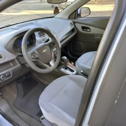 Interior do lado do motorista de um Chevrolet: volante, painel e bancos dianteiros de tecido cinza visíveis.