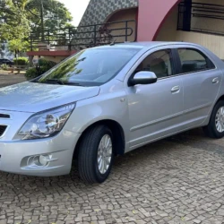 Sedã Chevrolet prateado estacionado em uma entrada pavimentada com pedras, com árvores e um prédio rosa ao fundo.
