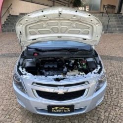 Vista frontal de um Chevrolet prateado com o capô aberto, expondo o compartimento do motor e a bateria em uma área externa pavimentada.
