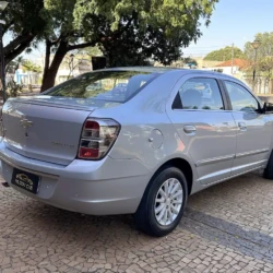 Vista de três quartos traseira de um sedã Chevrolet prateado estacionado sobre um piso de paralelepípedos, com árvores ao fundo e iluminação diurna.