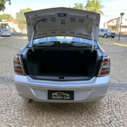 Porta-malas aberto de um sedã prateado, exibindo um espaço de carga amplo e vazio, em uma entrada de garagem.