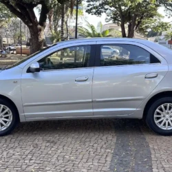 Sedã prateado de quatro portas estacionado em um pátio pavimentado com tijolos, com árvores ao fundo.