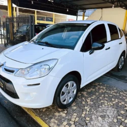 White compact hatchback car (Citroën) parked in a dealership lot under a covered area, front angled view, glossy finish