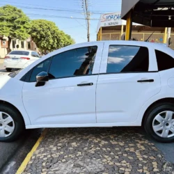 Side view of a white hatchback car parked along a curb on a sunny urban street with buildings in the background.