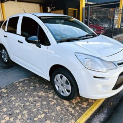 White hatchback car parked along a yellow-curb street with a tiled sidewalk and fence in the background, viewed from the front-left side.