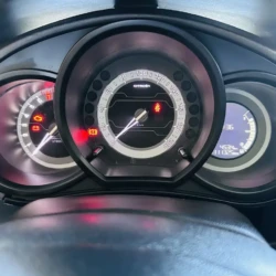 Car instrument cluster with illuminated warning lights, center speedometer, left tachometer, right digital/readout panel showing mileage and fuel.