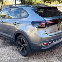 Rear three-quarter view of a metallic gray Volkswagen Nivus SUV parked on a cobblestone lot with roof rails and black alloy wheels on a sunny day, reflecting surroundings.